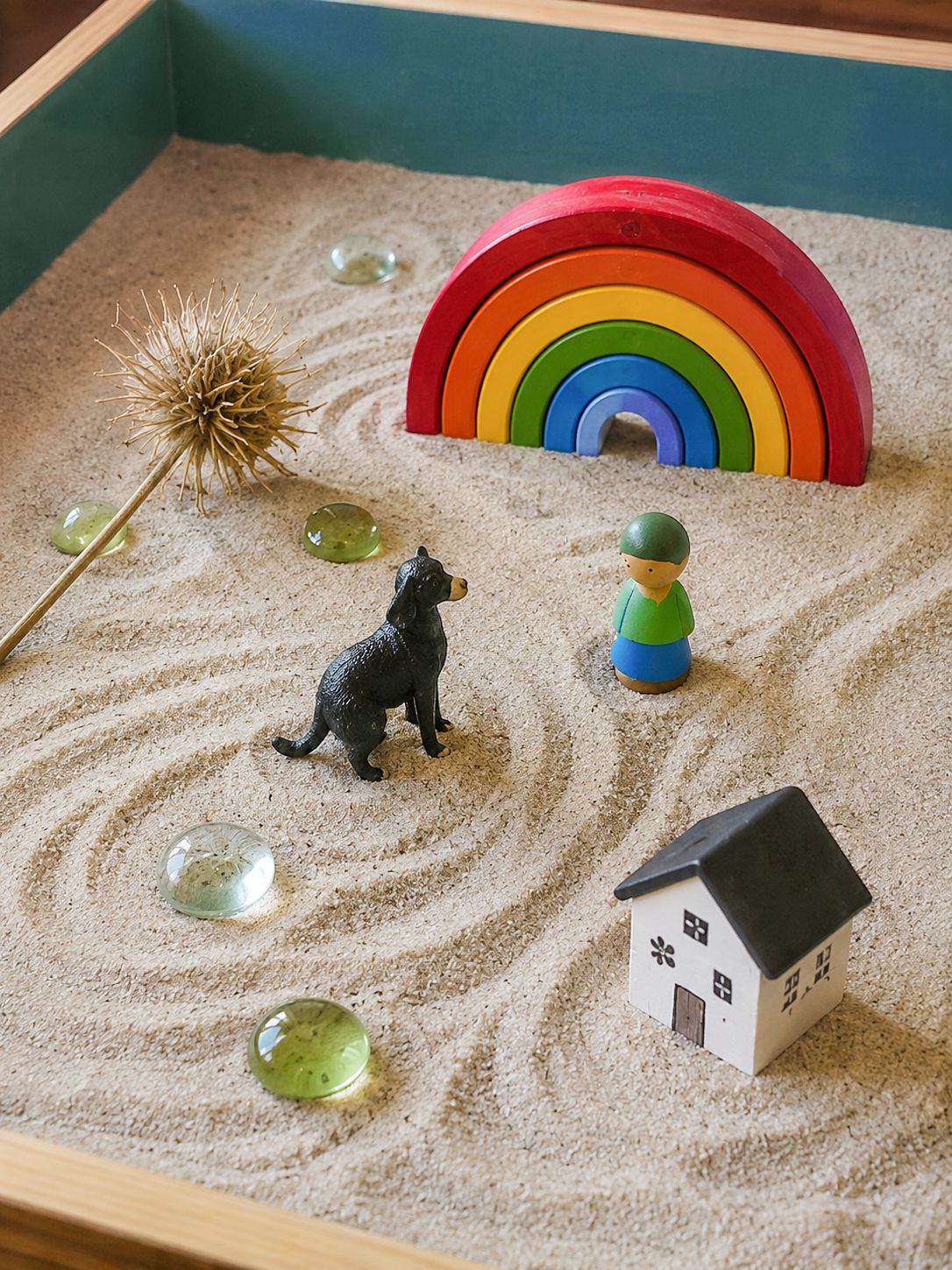 A sand tray with a small wooden rainbow, figures and a house arranged in raked sand